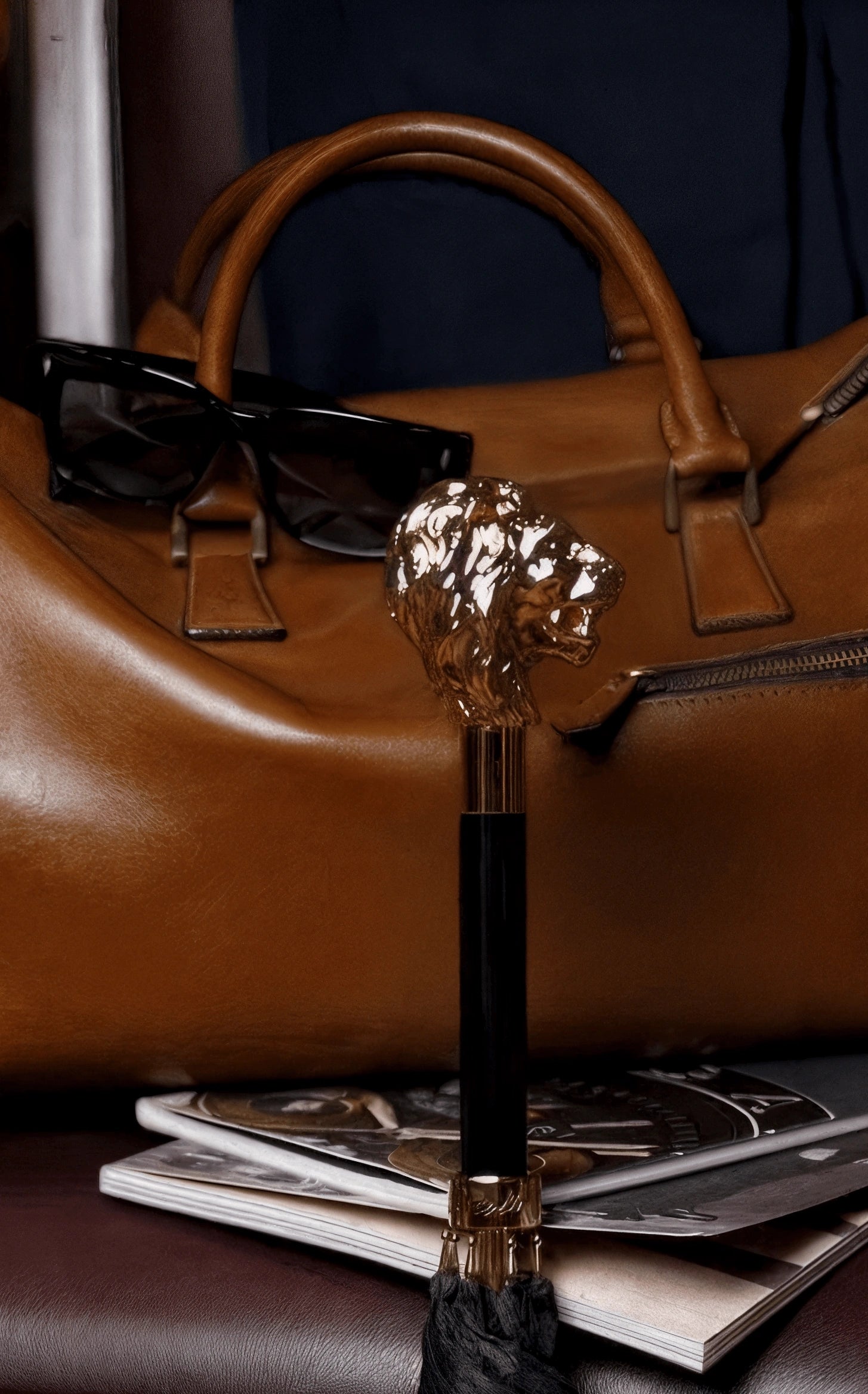 Brown leather handbag with sunglasses on top, gold and black decorative item in front, and magazines underneath.